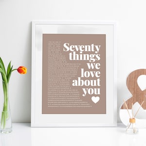 May include: A framed print with a brown background and white text that reads "Seventy things we love about you" with a heart symbol at the bottom. The print is sitting on a white surface with a wooden ampersand and a small glass bottle.