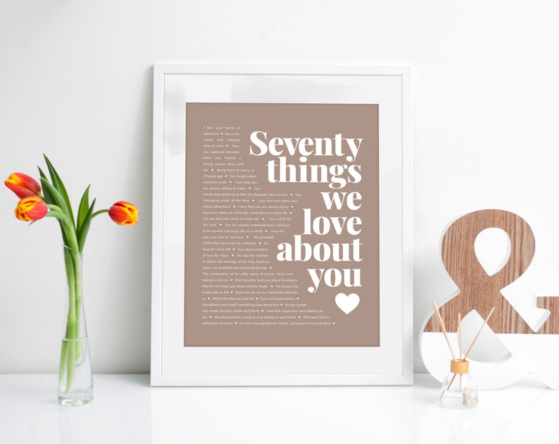 May include: A framed print with a brown background and white text that reads "Seventy things we love about you" with a heart symbol at the bottom. The print is sitting on a white surface with a wooden ampersand and a small glass bottle.