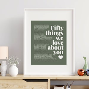 May include: A framed print with a green background and white text that reads "Fifty things we love about you" with a white heart at the bottom. The print is in a white frame and is sitting on a wooden shelf with other decorative items.