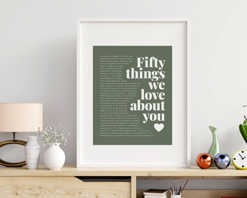 May include: A framed print with a green background and white text that reads "Fifty things we love about you" with a white heart at the bottom. The print is in a white frame and is sitting on a wooden shelf with other decorative items.