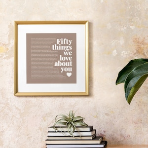 May include: A gold framed print with a brown background and white text that reads "Fifty things we love about you" with a small heart symbol at the bottom. The print is hanging on a textured wall.