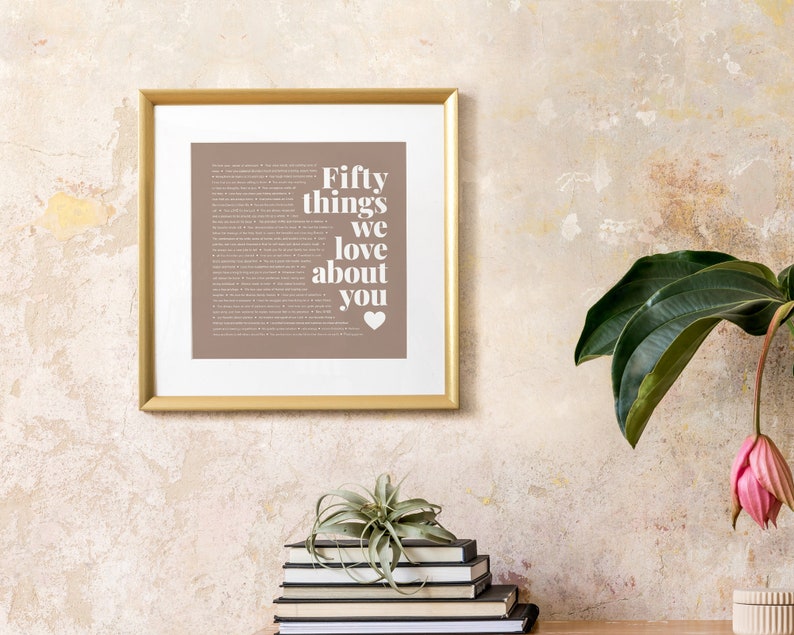 May include: A gold framed print with a brown background and white text that reads "Fifty things we love about you" with a small heart symbol at the bottom. The print is hanging on a textured wall.