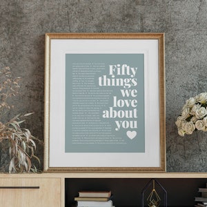 May include: A framed print with a blue background and white text that reads "Fifty things we love about you" with a white heart symbol. The print is in a gold frame and is sitting on a wooden shelf.