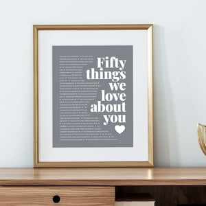 May include: A framed print with a gray background and white text that reads "Fifty things we love about you" with a heart symbol at the bottom. The print is in a gold frame and is sitting on a wooden table.