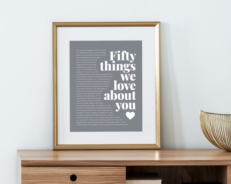 May include: A framed print with a gray background and white text that reads "Fifty things we love about you" with a heart symbol at the bottom. The print is in a gold frame and is sitting on a wooden table.