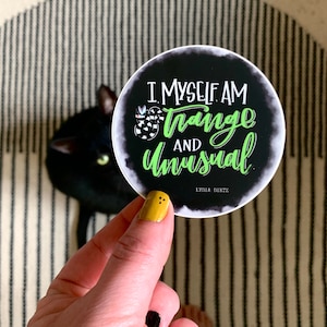 May include: A round black sticker with the text "I MYSELF AM Strange AND Unusual" in white and neon green. A black and white snake illustration is also on the sticker. The sticker is held in front of a black cat.