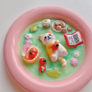 Pode incluir: Uma bandeja redonda rosa com uma cena fantasiosa com um gato branco e laranja segurando uma maçã. A cena inclui uma cesta de maçãs, biscoitos, um livro e outros alimentos em miniatura. O fundo é verde com pontos brancos.