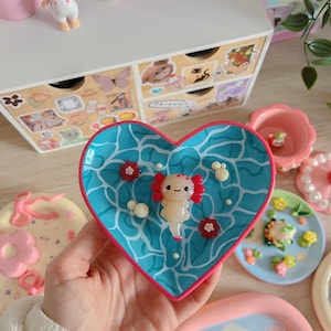 Op de afbeelding: Een hartvormig schaaltje met een blauw en wit waterontwerp, met een klein roze en wit axolotl-beeldje. Het schaaltje is omringd door andere decoratieve schaaltjes met verschillende ontwerpen.
