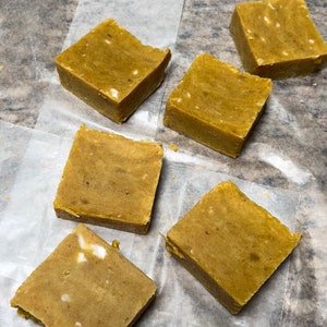 May include: Six bars of handmade soap, each with a light brown color and a speckled appearance. The soap bars are arranged on a white parchment paper.