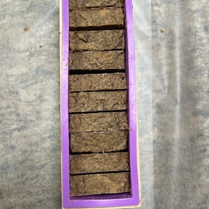 Può includere: Uno stampo in silicone viola riempito con 10 barre di sapone marrone. Il sapone ha un aspetto maculato.