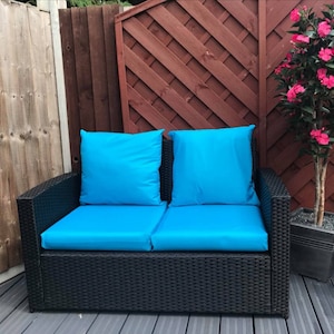 Op de afbeelding: Een zwarte wicker tweezitsbank met blauwe kussens. De bank staat op een houten terras met een houten hek en een potplant op de achtergrond.