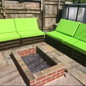 Op de afbeelding: Twee groene, gepolsterde palletsofa's met een bakstenen vuurplaats ertussen. De sofa's zijn gemaakt van houten pallets en hebben felgroene kussens.