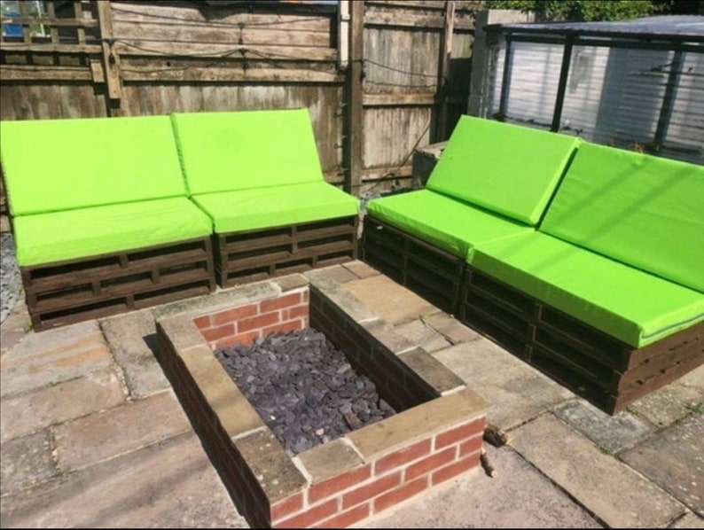 Op de afbeelding: Twee groene, gepolsterde palletsofa's met een bakstenen vuurplaats ertussen. De sofa's zijn gemaakt van houten pallets en hebben felgroene kussens.