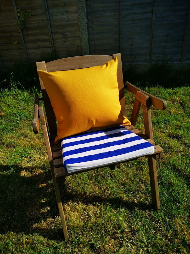 Op de afbeelding: Een houten stoel met een geel kussen en een blauw-wit gestreept kussen. De stoel staat op een grasveld.