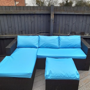 Op de afbeelding: Een blauwe hoekbank met een bijpassende poef. De bank heeft een zwart rieten frame en bestaat uit drie kussens. De poef is ook van zwart riet en heeft een blauw kussen.