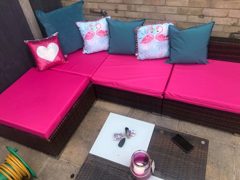 Op de afbeelding: Een roze rieten hoekbank met teal en witte kussens met patronen. De bank staat op een terras in de buitenlucht met een kleine tafel en een roze kaars.