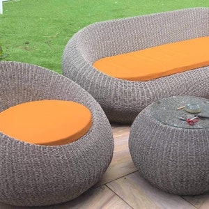 Op de afbeelding: Een wicker tuinmeubelset met een bank, twee stoelen en een ronde tafel. De meubels zijn grijs met oranje kussens.