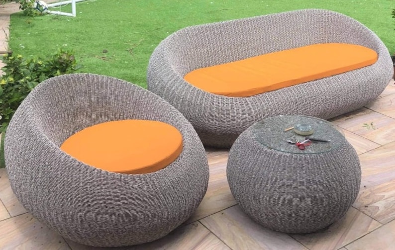 Op de afbeelding: Een wicker tuinmeubelset met een bank, twee stoelen en een ronde tafel. De meubels zijn grijs met oranje kussens.