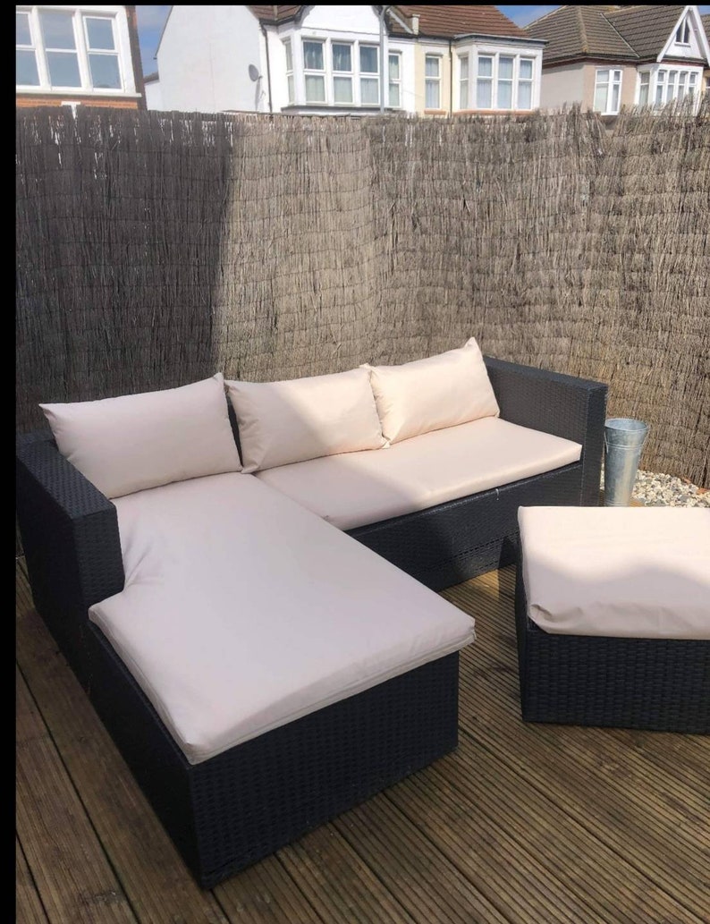 Op de afbeelding: Een zwarte wicker hoekbank met witte kussens. De bank heeft een chaise longue en een voetenbank. De bank staat op een houten terras.