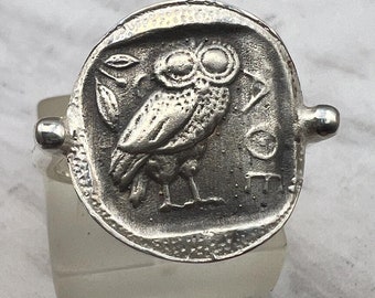 May include: A silver ring with an owl design. The owl is facing forward with its wings spread out. The ring is on a white marble surface.