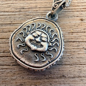 Pode incluir: Um colar com pingente de metal prateado apresentando um design de moeda circular com um rosto de sol sorridente em relevo no centro. O sol é cercado por raios curvos estilizados. O pingente está preso a uma corrente prateada com um passador decorativo.