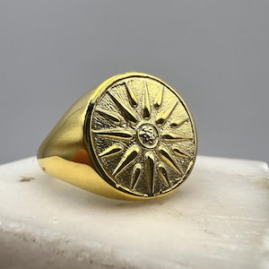 Könnte beinhalten: Ein goldener Ring mit einem runden, erhabenen Design einer Sonne mit 16 Strahlen. Der Ring liegt auf einer weißen Oberfläche.