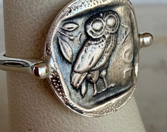 May include: A silver ring featuring a circular coin-style centerpiece with a raised relief of an owl and a sprig of leaves. The metal has a darkened, oxidized finish in the recessed areas, and the letters AOE are visible on the right side.