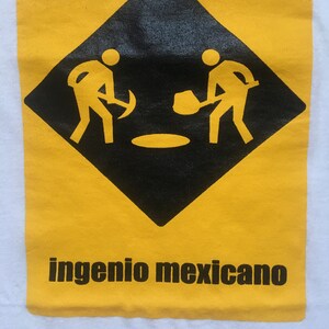 Könnte beinhalten: Weißes T-Shirt mit gelb-schwarzer Grafik. Die Grafik zeigt zwei stilisierte Figuren, die in einem schwarzen Diamanten graben. Der Text "ingenio mexicano" ist darunter in Schwarz gedruckt.