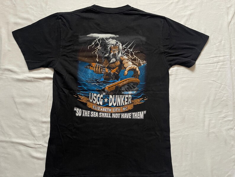 May include: Black t-shirt featuring a graphic of a bearded figure with a trident, lightning, and a boat. Text on the shirt reads "USCG DUNKER" and "SO THE SEA SHALL NOT HAVE THEM."