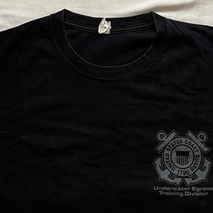 May include: Black t-shirt with a crew neck. The shirt features a gray graphic of the United States Coast Guard emblem on the right chest, with the text "Underwater Egress Training Division" below.