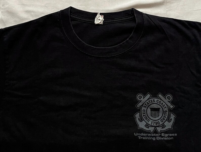 May include: Black t-shirt with a crew neck. The shirt features a gray graphic of the United States Coast Guard emblem on the right chest, with the text "Underwater Egress Training Division" below.