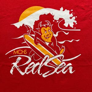 May include: Red t-shirt with a graphic of a devil surfing on a surfboard, with a yellow sun and white wave. The text "MCHS Red Sea" is printed in white.