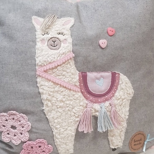 May include: A gray pillow with a white fluffy llama applique. The llama has pink accents, a pink saddle, and blue tassels. The pillow also has pink crocheted flowers and a "hand made" tag.