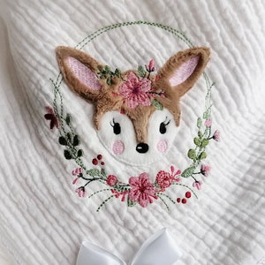 Könnte beinhalten: Weiße Musselin-Babydecke mit einem zarten gestickten Design eines Rehs mit einem Blumenkranz. Die Decke hat eine weiße Satinschleife und einen weißen Häkelrand.