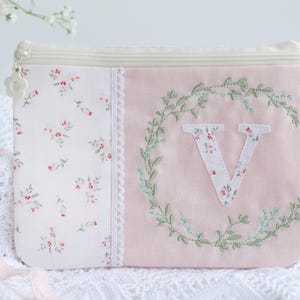 Op de afbeelding: Een rechthoekig, roze en wit bloemenprint etui met ritssluiting. De etui heeft een bloemenkrans met de letter "V" in het midden. De ritstrekker is een witte bloem. Afmetingen in centimeters.