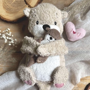 Könnte beinhalten: Ein beiges und weißes Plüschotter-Spielzeug, das einen kleineren Otter umarmt. Das Spielzeug hat eine schwarze Nase und rosa Pfoten. Ein rosa Filzherz mit gesticktem "I U" liegt in der Nähe. Die Szene spielt sich auf einem neutralen Leinentuch ab.