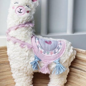 Puede incluir: Juguete de peluche de llama blanco con detalles en rosa y azul. La llama tiene un coraz&oacute;n rosa en la espalda y una nariz rosa.