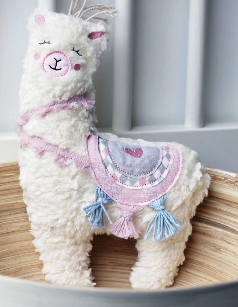 Puede incluir: Juguete de peluche de llama blanco con detalles en rosa y azul. La llama tiene un coraz&oacute;n rosa en la espalda y una nariz rosa.