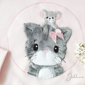 May include: A pink sweatshirt with a gray cat and a gray mouse embroidered on it. The cat has a pink bow on its head. The text "Johllina" is embroidered on the bottom right corner of the sweatshirt.