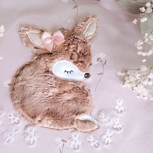 May include: A brown and white embroidered fawn with a pink bow sleeping on a pink background with white embroidered flowers.