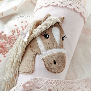 Pode incluir: Um artigo cilíndrico de tecido rosa com uma aplicação de cabeça de cavalo. O cavalo tem uma crina trançada, uma rédea branca e um pequeno laço branco. O artigo é adornado com renda e tem o nome "Johlina" em itálico.