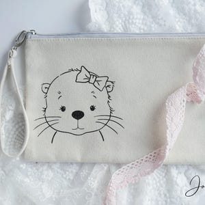 Könnte beinhalten: Eine weiße Canvas-Reißverschlusstasche mit einer schwarz-weißen Illustration eines Otters mit Schleife. Die Tasche hat einen rosa Spitzenbesatz und einen schwarzen Riemen.