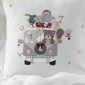 May include: White cushion with an embroidered design of a grey van laden with Christmas gifts, a tree, and soft toys. A rabbit and a bear sit in the front. The van has a wreath and is decorated with lights and stars.