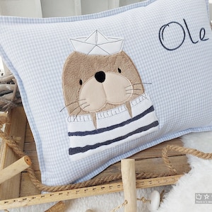 Könnte beinhalten: Ein hellblaues Kissen mit weißem Gingham-Muster zeigt ein gesticktes Bild eines braunen Seehundes, der einen Matrosenhut und einen gestreiften Pullover trägt. Der Name "Ole" ist in Schwarz auf das Kissen gestickt.