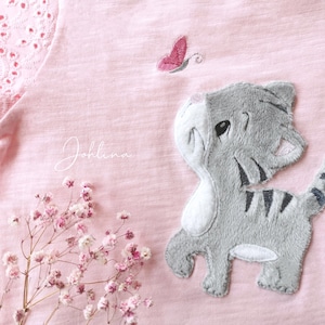 Op de afbeelding: Een roze T-shirt met een geborduurd grijs katje dat omhoog kijkt naar een roze vlinder. Het shirt heeft een witte kantversiering op de schouder.