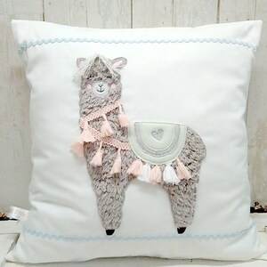 May include: A white pillow with a light blue trim features a gray and pink embroidered llama with tassels. The llama has a heart on its back.