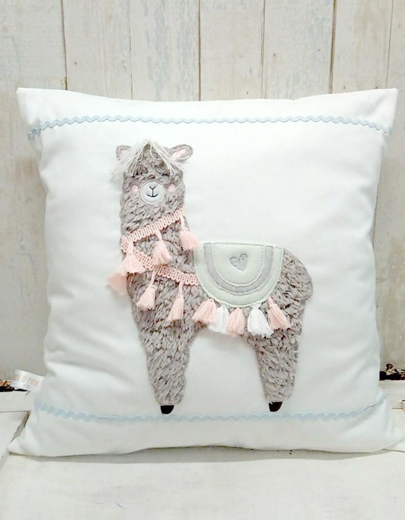 May include: A white pillow with a light blue trim features a gray and pink embroidered llama with tassels. The llama has a heart on its back.