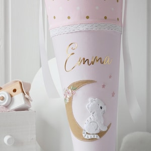 Könnte beinhalten: Rosa-goldener Schultüte mit einer weißen Katze, die auf einem goldenen Halbmond sitzt. Die Schultüte hat weiße Punkte und eine weiße Spitzenborte. Der Name "Emma" ist in goldenen Buchstaben auf der Schultüte geschrieben.