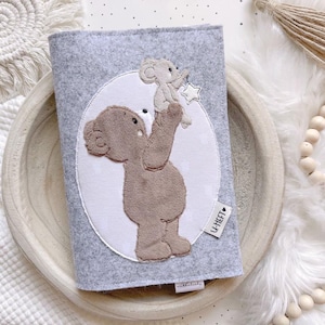 Puede incluir: Una funda de libro de fieltro gris con un oso aplicado que sostiene un oso más pequeño y una estrella. El diseño está dentro de un óvalo blanco. La funda tiene una pequeña etiqueta que dice "U-HEFT".
