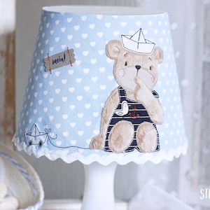 Puede incluir: Una pantalla de lámpara azul con corazones blancos y un oso de peluche marrón que lleva un traje de marinero. El oso de peluche sostiene un pequeño pájaro blanco y tiene un barco de papel en la cabeza. La pantalla de la lámpara tiene un borde festoneado y una pequeña etiqueta que dice "mein!"